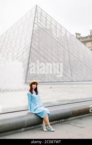 PARIS, FRANCE - 17 septembre 2019 : jeune femme heureuse en robe bleue, assise près de la fontaine et posant pour une photo avec la pyramide de verre Banque D'Images
