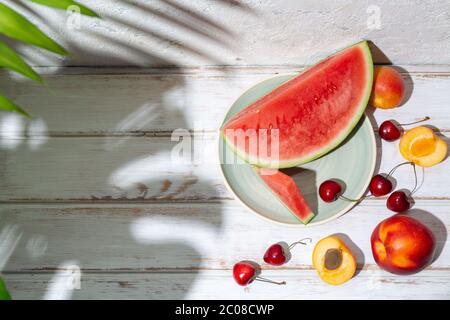 Composition de fruits d'été avec ombres de feuilles tropicales sur une table en bois blanc et un mur en béton. Copier l'espace Banque D'Images