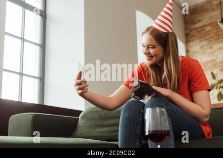 objectif sélectif de jeune femme attrayante dans la tête de fête prenant selfie avec chat près d'un verre de vin rouge Banque D'Images