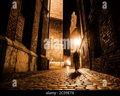 Rue pavée éclairée avec réflexions claires sur les pavés de la vieille ville historique la nuit. La silhouette sombre floue de la personne évoque Jack l'Éventreur. Banque D'Images