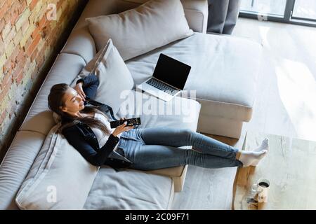 vue en hauteur de la femme heureuse tenant la télécommande près de l'ordinateur portable avec écran vierge sur le canapé et une tasse sur la table basse Banque D'Images