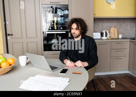 Attention sélective de l'homme écrivant sur des notes adhésives pendant le séminaire en ligne sur ordinateur portable dans la cuisine Banque D'Images