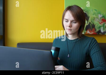 Une fille au visage calme s'assoit à un ordinateur portable à une table dans un café, sur un canapé marron, contre un mur jaune. Il regarde un moniteur d'ordinateur, tenant une tasse de café réutilisable écologique dans sa main. Banque D'Images