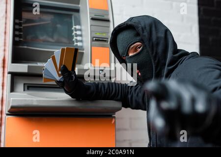 Foyer sélectif de cambrioleur dans le balaclava pointant avec le doigt à la caméra tout en tenant des cartes de crédit près de atm Banque D'Images