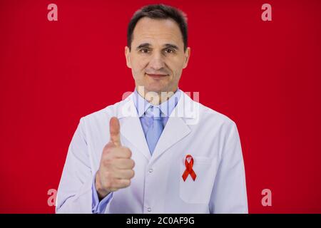Un médecin de grand âge souriant, portant un manteau blanc et un ruban rouge, montre un signe Banque D'Images