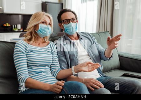 Foyer sélectif de la femme dans le masque médical regardant la télévision près de mari pointant avec la main sur le canapé Banque D'Images