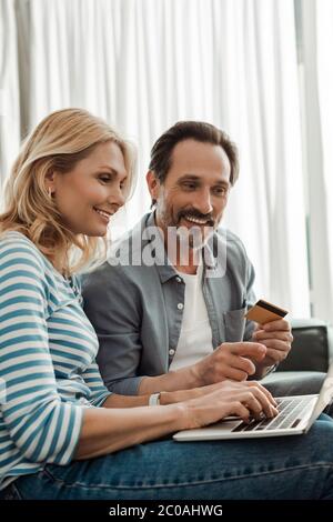 Femme souriante utilisant un ordinateur portable près de mari tenant une carte de crédit et pointant avec le doigt à la maison Banque D'Images