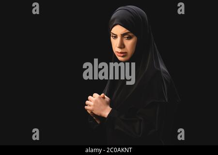 Vue latérale de la femme musulmane contrariée dans le hijab avec les mains serrées isolées sur le noir Banque D'Images