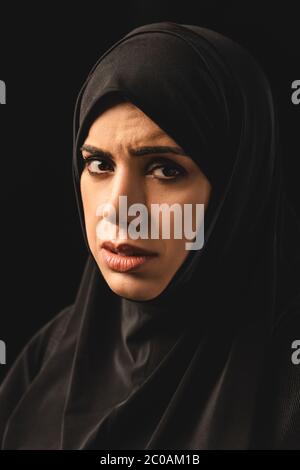 Portrait de la femme musulmane bouleversée dans le hijab regardant la caméra isolée sur noir Banque D'Images