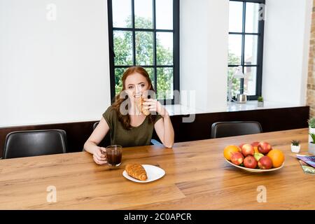 Une femme aux cheveux rouges mangeant un croissant en tenant une tasse de café à la maison Banque D'Images