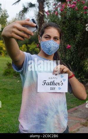 Jeune fille souriant au téléphone portable pendant un appel vidéo à un père à la maison pour la fête des pères. Elle tient une carte blanche avec le texte Happy Pather's Day Banque D'Images