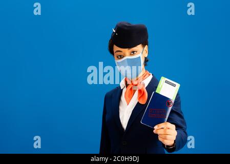 un agent de bord afro-américain dans un masque médical tenant un passeport et un billet d'avion isolés en bleu Banque D'Images