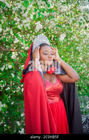 Reine dans la couronne et le manteau royal. Jolie fille sur le fond d'un jardin fleuri. Afro-américain en rouge Banque D'Images