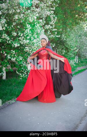 Reine dans la couronne et le manteau royal. Jolie fille sur le fond d'un jardin fleuri. Afro-américain en rouge Banque D'Images