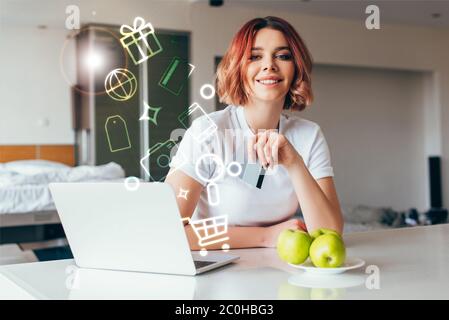 sourire fille shopping en ligne avec ordinateur portable et cartes de crédit et des signes de shopping sur la cuisine avec des pommes pendant l'auto-isolation Banque D'Images