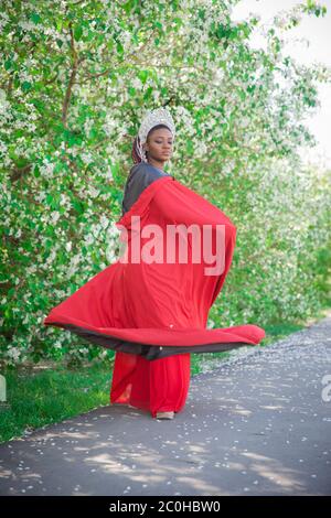 Reine dans la couronne et le manteau royal. Jolie fille sur le fond d'un jardin fleuri. Afro-américain en rouge Banque D'Images