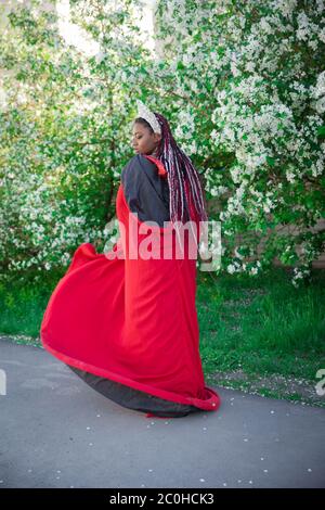 Reine dans la couronne et le manteau royal. Jolie fille sur le fond d'un jardin fleuri. Afro-américain en rouge Banque D'Images