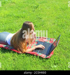 Une belle jeune fille blanche dans un T-shirt blanc et avec de longs cheveux se trouve sur un tissu rouge, sur l'herbe verte, sur la pelouse et de travail derrière un ordinateur portable noir. Banque D'Images