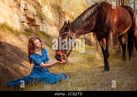 Belle fille dans une robe bleue nourrit un cheval avec une pomme de ses mains. Photographie de conte de fées, artistique, magique. Fille et cheval dans le coucher du soleil Banque D'Images