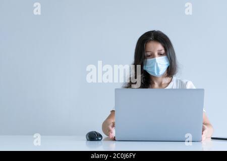 La fille travaille de la maison pendant la quarantaine. Pandémie du virus Corona. Une fille dans un masque chirurgical travaille sur un ordinateur portable et boit du café. Travail à distance. Banque D'Images