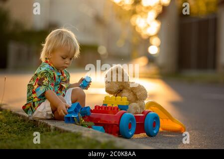 Beau tout-petit enfant, jouant avec des jouets en plastique, des blocs, des voitures au coucher du soleil dans un petit village Banque D'Images