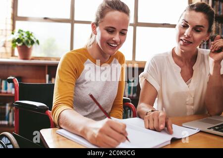 Une adolescente assise dans un fauteuil roulant écrivant dans un carnet avec une enseignante assise à l'école. Femme handicapée étudiant avec un tuteur. Banque D'Images