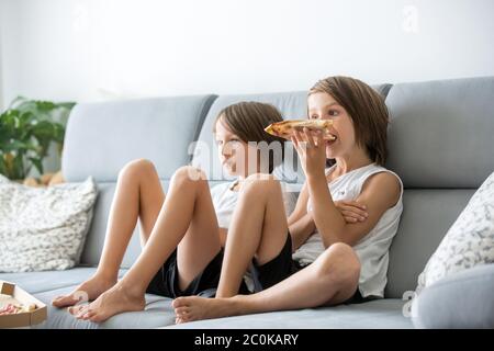 Enfants adorables, assis sur un canapé, manger de la pizza et regarder la télévision. Enfant affamé prenant un morceau de pizza lors d'une fête à la maison Banque D'Images