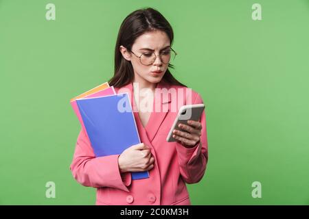 Portrait d'une belle jeune femme pensive élégante portant une veste rose debout isolée sur fond vert, à l'aide d'un téléphone portable, dossier de maintien Banque D'Images