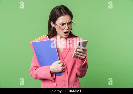 Portrait d'une belle jeune femme élégante et confuse portant une veste rose sur fond vert, isolée à l'aide d'un téléphone portable, tenant un dossier Banque D'Images