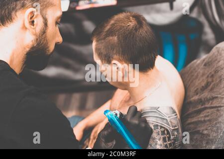 Salon de tatouage. Le maître de tatouage tatoue un homme sur son épaule. Machine de tatouage, sécurité et hygiène au travail. Gros plan, teinté, tatoueur Banque D'Images