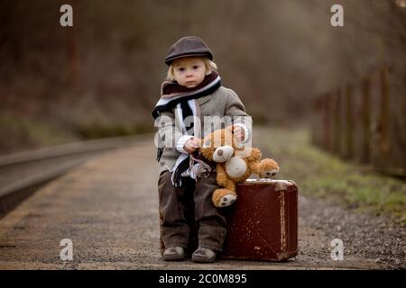 Adorable garçon dans une gare, attendant le train avec une valise et un jouet ours en peluche Banque D'Images