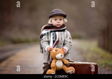 Adorable garçon dans une gare, attendant le train avec une valise et un jouet ours en peluche Banque D'Images