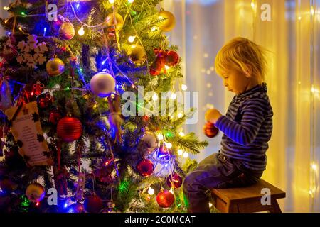 Beau garçon blond, décorant l'arbre de noël avec des boules et des cordes légères à la maison Banque D'Images