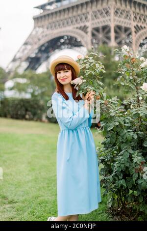 Belle jeune femme à cheveux rouges, vêtue d'une robe élégante bleue et d'un élégant chapeau de paille, posant près des rosiers fleuris sur la vieille ville européenne de Paris Banque D'Images