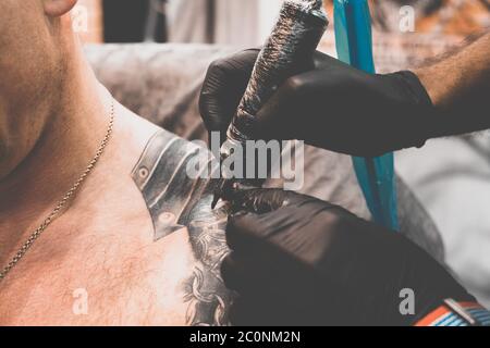 Salon de tatouage. Le maître de tatouage tatoue un homme sur son épaule. Machine de tatouage, sécurité et hygiène au travail. Gros plan, teinté, tatoueur Banque D'Images