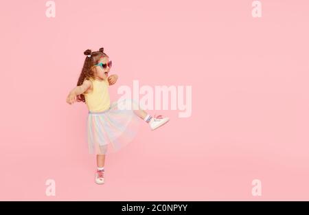 Portrait de petite fille surprise dans des lunettes de soleil sur fond rose. Modèle enfant ont plaisir et saut. Publicité des produits pour enfants Banque D'Images