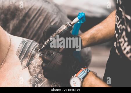 Salon de tatouage. Le maître de tatouage tatoue un homme sur son épaule. Machine de tatouage, sécurité et hygiène au travail. Gros plan, teinté, tatoueur Banque D'Images