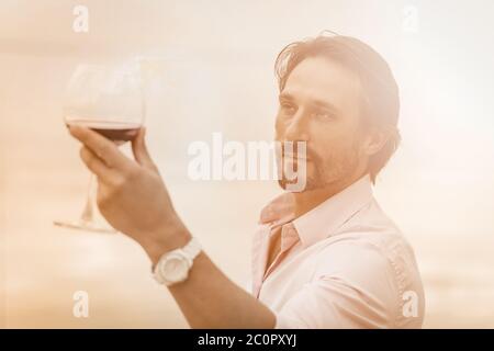 L'homme caucasien tient un verre de vin rouge. Silhouette masculine avec lunettes de soleil dans le dos éclairé. Concept de dégustation de vins. Image en tons Banque D'Images