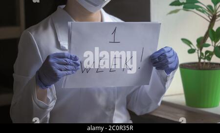 Le médecin en uniforme blanc montre une inscription d'une semaine sur un morceau de papier. Femme debout près de la fenêtre dans le bureau médical. Concept de quarantaine Banque D'Images