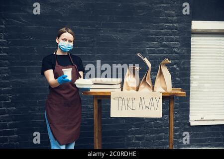 Protégé. Femme préparant des boissons et des repas, portant un masque facial et des gants de protection. Service de livraison sans contact pendant la pandémie de coronavirus de quarantaine. Concept à emporter. Mugs à câble recycable, paquets. Banque D'Images