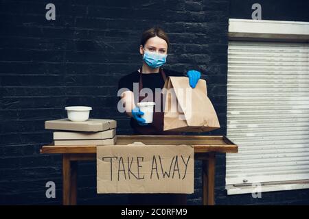 Choisissez la santé. Femme préparant des boissons et des repas, portant un masque facial de protection, des gants. Service de livraison sans contact pendant la pandémie de coronavirus de quarantaine. Concept à emporter. Mugs à câble recycable, paquets. Banque D'Images