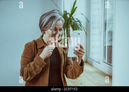 Une femme joyeuse aux cheveux argentés communique en ligne sur une tablette numérique. Belle femme mûre sourit avoir une réunion en ligne avec ses gens adorables qui séjournent Banque D'Images