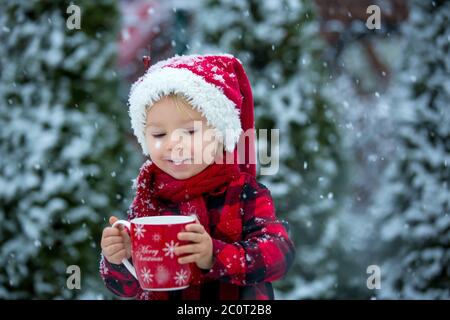 Joli petit garçon, tenant une tasse de lait chaud, buvant dans la neige en plein air, profitant des vacances d'hiver et de Noël Banque D'Images