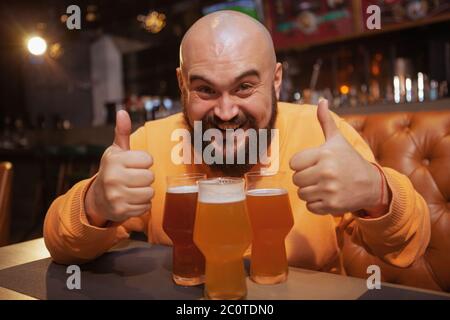 Un homme à barbe qui rit, se fait un plaisir de montrer ses pouces assis devant trois verres à bière au pub Banque D'Images