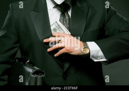 Homme d'affaires met une main de billet de banque dans sa poche tenant une serviette noire sous son bras. Homme caucasien portant une combinaison noire tenant de l'argent sur le gris Banque D'Images
