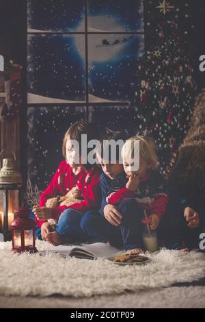 Trois enfants, frères garçons, lisant un livre à la nuit de Noël dans une chambre confortable aux bougies Banque D'Images