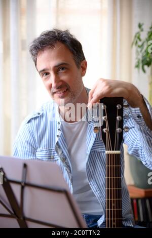 Gros plan d'un homme souriant, appuyé sur le chevalet d'une guitare, regardant l'appareil photo et le support musical avec une séparation devant la maison. Banque D'Images