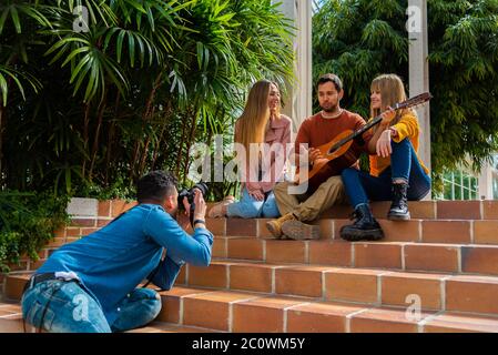 Un musicien avec une guitare et deux jeunes filles posent sur un escalier à côté de quelques arbres pour un photographe, un espace pour le texte et la lumière naturelle Banque D'Images