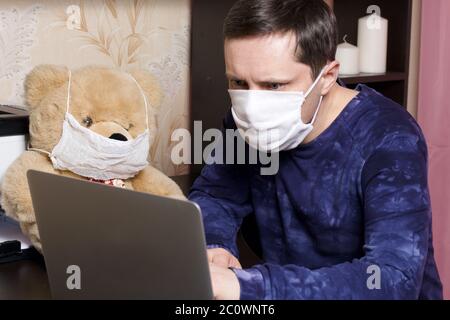Un homme dans un masque de gaze de protection travaille derrière un ordinateur portable dans des conditions d'auto-isolation. À proximité, vous trouverez un jouet. Porter un masque de gaze de protection. Banque D'Images