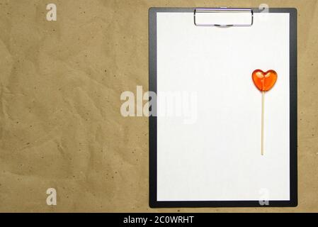 La tablette avec un clip pour le clip de papier avec une feuille blanche a4 repose sur l'arrière-plan du papier brun froissé. Sur le dessus se trouve un lollipop de caramel de rouge orange en forme de coeur. Place pour le texte et la mise en page pour la conception. Banque D'Images